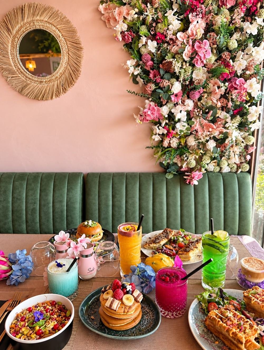 Une table de brunch dressée devant le mur fleuri, la banquette en velours vert et le miroir en rotin